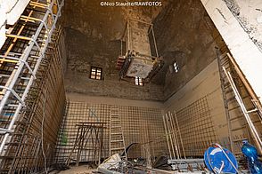 Blick von unten nach oben in eine große historische Halle. Rathauses mit der schwebenden Säule. Eine Säule wurde mit Holz und Metallverstrebungen stabilisiert und schwebt im Raum. Die Wände im Kellerbereich sind mit Baustahlmatten ausgekleidet.
