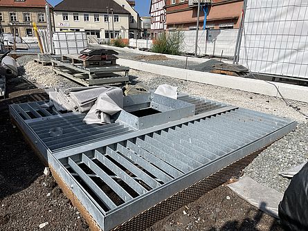Auf einer großen Baustelle sieht man eine Metallvorrichtung im Boden eingelassen. Im Umfeld ist eine Baustelle mit Absperrungen und Baumaterial. Am Rand der Baustelle sieht man Häuser.