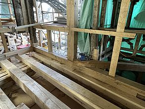 Auf einer Baustelle sieht man Zimmererarbeiten. Am Boden liegen neue Holzbalken. Auch das Holzgerüst mit den Stützen der Außenwände wurden erneuert. Das Mauerwerk wurde noch nicht zwischen den Balken eingezogen. Der Blick geht nach Außen und zeigen ein Außengerüst und grüne Abdeckplanen.