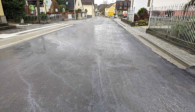 Eine frisch geteerte Straße. Links und rechts befinden sich Wohnhäuser.