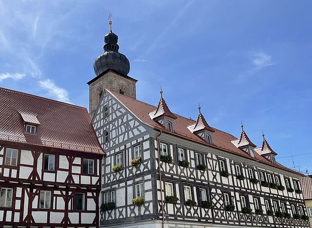 Mehrere Fachwerkhäuser reihen sich aneinander. Im Hintergrund ragt ein Kirchturm auf. Darüber strahlt ein blauer Himmel.
