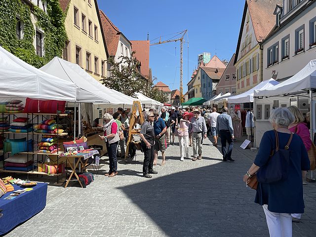 Eine Straße mit weißen Pavillons und Marktständen. Menschen schlendern auf der Straße und betrachten die Auslagen. Rechts und links sieht man die Häuserfronten.