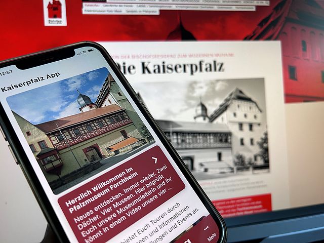 Im Vordergrund befindet sich ein Handy, das eine App zeigt. Auf dem Display sieht man die Frontansicht einer Burg und unterhalb einen rot hinterlegten Text. Im Hintergrund sieht man verschwommen einen Computerbildschirm.