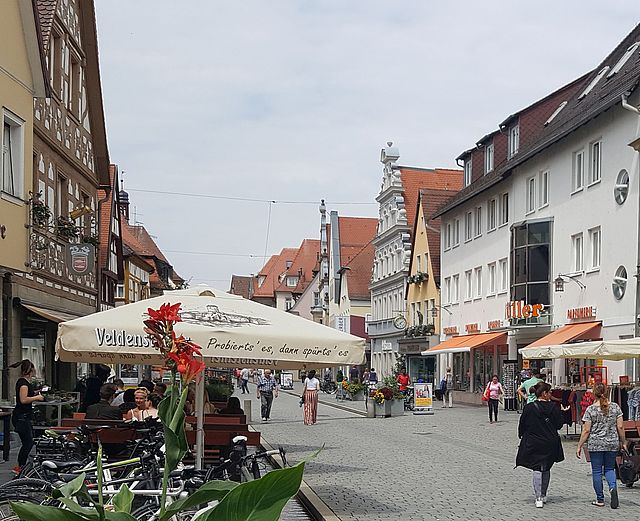 Passanten bummeln durch die Forchheimer Fußgängerzone. Man sieht die Ladenzeile der Hauptstraße und Restaurantgäste, die auf Bänken an Tischen sitzen. Links vorne blüht eine rote Sommerpflanze, dahinter stehen Räder.