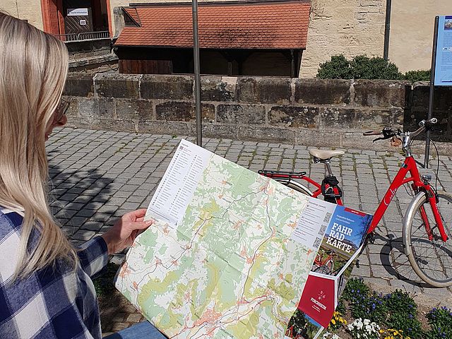 Eine blonde Frau betrachtet die Touren in der neuen Radkarte. im Hintergrund sieht man ein rotes Damenrad und den Eingangsbereich mit Tor der Kaiserpfalz.