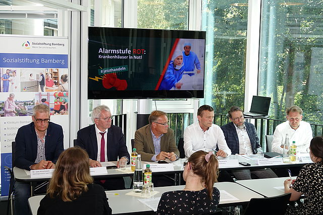Sechs Vertreter der Städte und Kliniken Bamberg und Forchheim sowie der Landrat des Landkreises Bamberg sitzen bei einer Pressekonferenz vor Medienvertretern. Im Hintergrund zeigt ein Bildschirm zwei Krankenpfleger im Einsatz sowie den Schriftzug "Alarmstufe ROT: Krankenhäuser in Not!" auf schwarzen Untergrund und ein rotes Megafon sowie den Hashtag #jetzthandeln