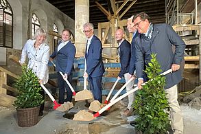 Fünf Männer und eine Frau halten jeweils eine mit Sand gefüllte Schaufel in die Mitte. Sie befinden sich in einer großen Halle, in der Baugerüste und Holzstützen zu sehen sind.