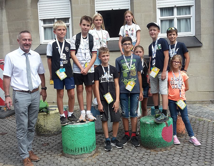 Kinder im Schulalter stehen in einer Gruppe mit Bürgermeister Udo Schönfelder vor dem Verwaltungsgebäude. Einige Kinder tragen Schirmmützen und rote Jutebeutel mit der Aufschrift "Entdecker Täschla".