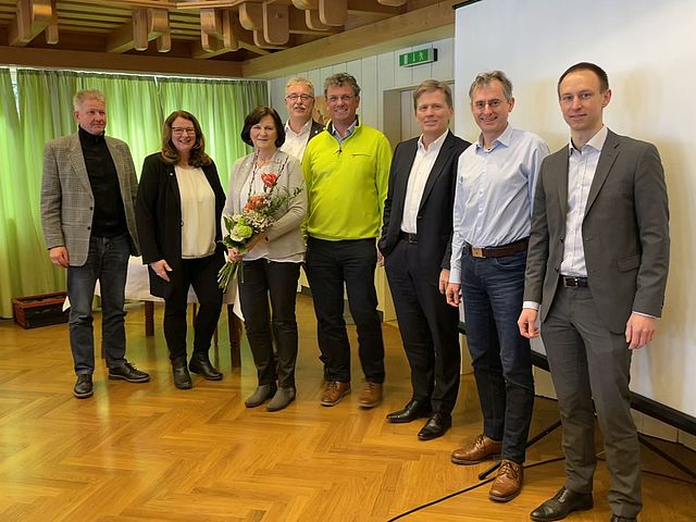 Acht Vertreterinnen und Vertreter der Arbeitsgemeinschaft der Oberfränkischen Wohnungsunternehmen stehen in einer Reihe vor einer weißen Wand und einer Leinwand. Eine Dame hält einen Blumenstrauß in den Händen.