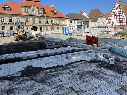 Ein großer Platz in einer Stadt wird neu gestaltet. Man sieht eine große Baustelle. Pflasterarbeiten mit hellgrauen Steinen wurden schon fertiggestellt. Einige Paletten mit Pflastersteinen, Baumaterial und ein Bagger sind zu sehen. Der Platz ist von Häusern umgeben.