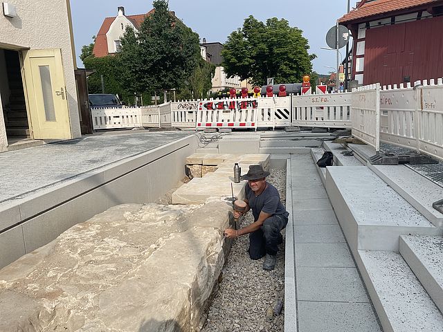 Ein Steinmetz bearbeitet die Sandsteinquader der freigelegten alten Stadtmauer. Neu gepflasterte Wege und Stufen umgeben die Mauersteine. Seitlich und hinten sichern Absperrgitter die Baustelle.