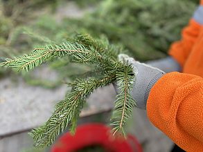 Man sieht zwei mit Arbeitshandschuhen geschützte Hände, die frische Tannenzweige vor einem Arbeitstisch bearbeiten.