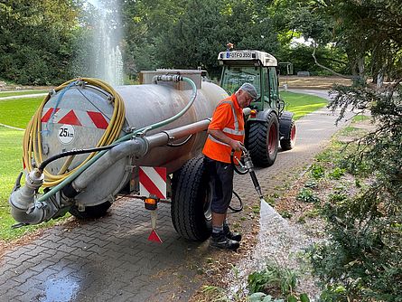 Ein städtischer Mitarbeiter gießt mit Hilfe eines großen Wassertanks Beete im Stadtpark. Im Hintergrund sieht man einen Springbrunnen und hohe Bäume.