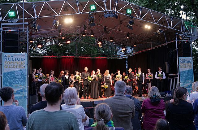 Konzert des KulturSommerQuartiers in Forchheim. Auf einer Bühne steht das Ensemble des Symphony Orchestra mit Blumensträußen in den Händen.