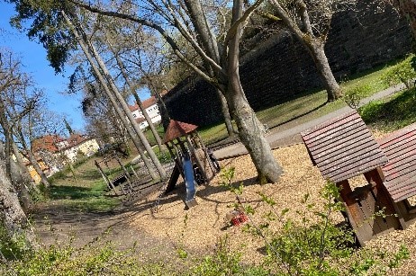 Ein Spielplatz in einem Park an einer großen Stadtmauer mit Rutschenturm, Spielhaus und Seilbahn.