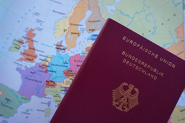 Man sieht im rechten Vordergrund einen dunkelroten Deutschen Reisepass mit goldenem Text. Der Text lautet "Europäische Union - Bundesrepublik Deutschland". Darunter befindet sich der Reichsadler, das deutsche Wappen der Bundesrepublik Deutschland. Im Hintergrund sieht man eine bunte Europakarte.