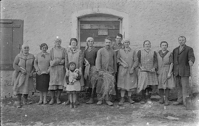 Schwarzweiß-Aufnahme von Personen in ländlicher Kleidung vor einem Gebäude.