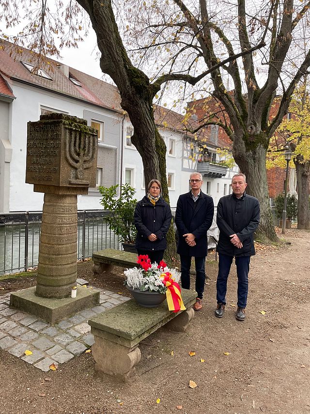 Zwei Männer und eine Frau stehen an einem Synagogendenkmal. Vor ihnen steht eine bepflanzte Blumenschale.