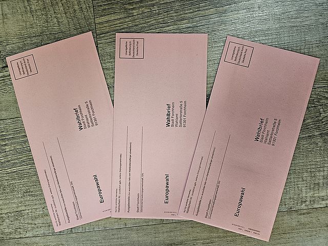 Drei rote Wahlbriefumschläge mit dem Aufdruck Europawahl liegen aufgefächert auf einem Holztisch.