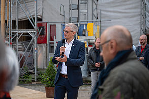 Ein Mann hält eine Rede. Im Vordergrund sieht man verschwommen Menschen. Im Hintergrund befindet sich eine Baustelle.