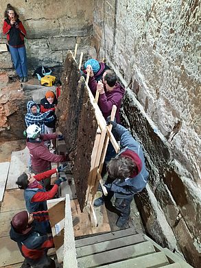 Mehrere Arbeiter transportieren das Lackprofil eine Treppe hinauf aus dem Kellerbereich des historischen Rathaus.