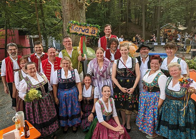 Eine große Gruppen Männer und Frauen in fränkischer Tracht befinden sich auf einem Walgelände mit Biergarten. Im Hintergrund sieht man Besucher und eine Essensbude.