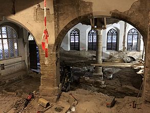 Blick von oben auf Ausgrabungen in einer großen historischen Halle. Die Halle wird von großen Steinsäulen gestützt. Man sieht Werkzeuge und einen Bagger in der Halle verteilt.