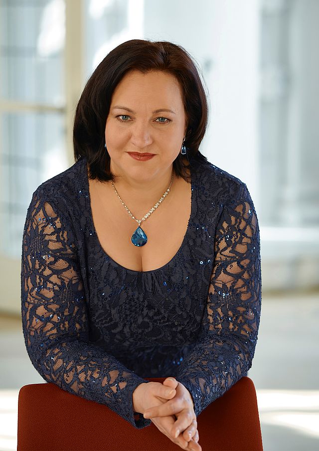 Die Preisträgerin Elena Belakova stützt sich mit den Unterarmen auf einen roten Stuhl. Sie trägt ein schwarzes Spitzenoberteil und eine Halskette. Die Frau lächelt in die Kamera.