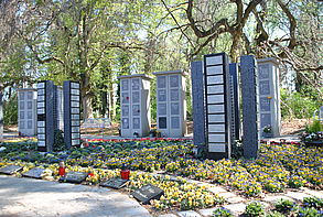 Man sieht im Vordergrund ein großes Beet mit Frühlingsblumen, die Friedhofssteelen umrahmen. Im Hintergrund stehen große Bäume.