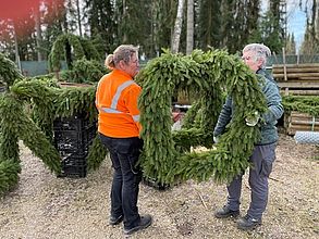 Zwei Frauen in Arbeitskleidung tragen eine mit Tannenzweigen geschmückte Krone für den Osterbrunnen.