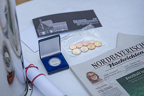 Auf einen weißen runden Tische befindet sich links eine silbrige Edelstahlkapsel. Auf dem Tisch befinden sich zwei Tageszeitungen, Euro-Münzen, zwei Einladungen in schwarzweiß-Druck, gerollte Baupläne, ein Geburtstaler der Stadt Forchheim, ein Flyer der Tourist-Information und die Verschlusskappe der Edelstahl-Kapsel.