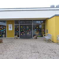 Bunt beklebte Glastüren und Eingangsbereich der Kita Sattlertor mit der gelben Außenfassade und zwei Metall-Sitzbänken am Eingang.
