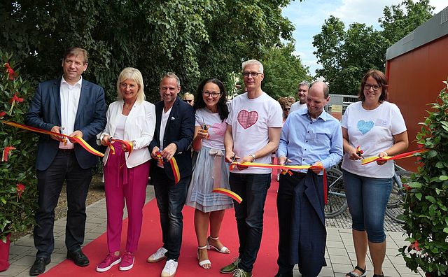 Vertreterinnen und Vertreter der Stadt Forchheim und Politik sowie die Leitungen der KiTa Sattlertor und Sattlertörchen durchschneiden das rot-gelbe Band mit den Farben der Stadt Forchheim zur Einweihung der neuen Kita.