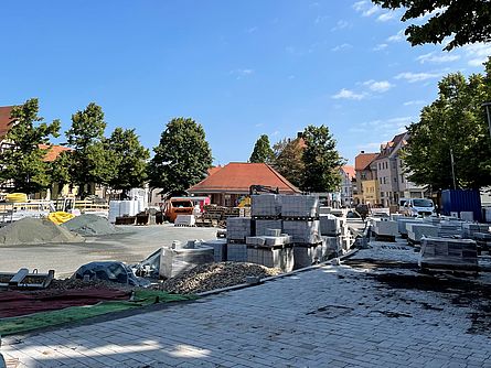Ein großer Platz in einer Stadt wird neu gestaltet. Man sieht eine große Baustelle. Pflasterarbeiten mit hellgrauen Steinen wurden schon fertiggestellt. Einige Paletten mit Pflastersteinen, Baumaterial, Baugeräte sind zu sehen. Der Platz ist von großen Bäumen umgeben. In der Mitte steht ein Haus mit rotem Dach.