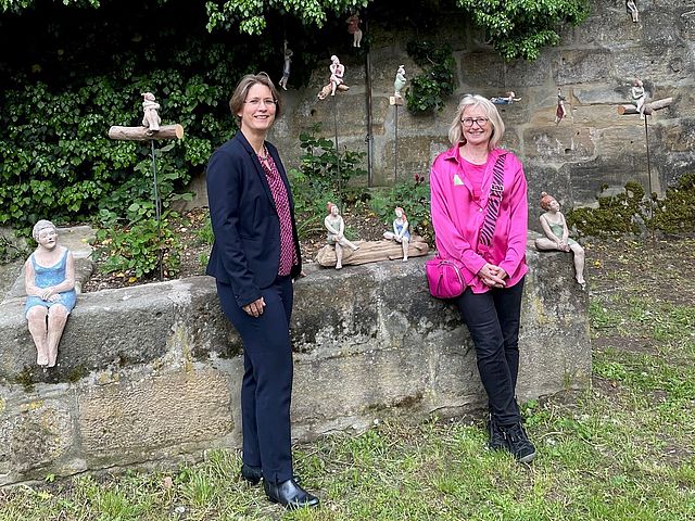 Bürgermeisterin Dr. Annette Prechtel trägt einen dunkelblauen Anzug und eine lilafarbene Bluse. Sie steht vor einer Sandsteinmauer und einem Hochbeet. Auf dem Rand sitzen vier getöpferte kleine Damenfiguren in kurzen Kleidern und verschiedenen Sitzpositionen. Rechts im Bild lehnt Museumsleiterin Susanne Fischer am Hochbeet. Sie trägt einen schwarze Hose und eine pinkfarbene Jacke und eine farblich angepasste Umhängetasche. Beide lächeln in die Kamera.
