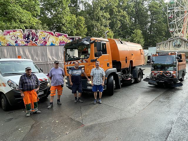 Vier Männer in Arbeitskleidung stehen vor einer großen und kleinen Kehrmaschine sowie einem kleinen Tieflader auf einem Festgelände mit Buden und Fahrgeschäften.