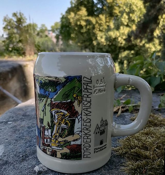 Das neue Steingut Annafest-Krügla 2023 steht auf einem Sandstein. Ein farbiges Motiv mit einer Musikkapelle am Festgelände im Kellerwald ziert den Krug. Rechts befindet sich ein Schriftzug "Förderkreis Kaiserpfalz". Rechts daneben sieht man das Logo des Pfalzmuseum Forchheim. Im Hintergrund befinden sich große Bäume und Sträucher.