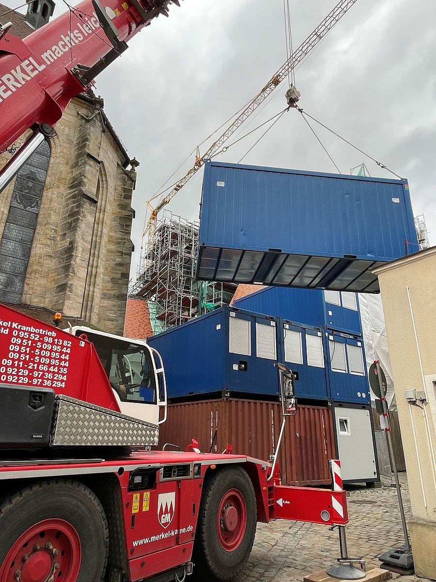 Die neuen Container für die Haustechnikgewerke an der Rathausbaustelle werden gesetzt. Ein Kran stapelt große Baucontainer übereinander.
