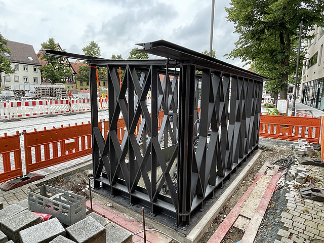 Ein moderner Hochbau aus dunklem Stahl wird gerade an einem Treppenabgang zu einer Tiefgarage errichtet. Umliegend befindet sich eine Großbaustelle mit Absperrungen und verschiedenem Baumaterial.