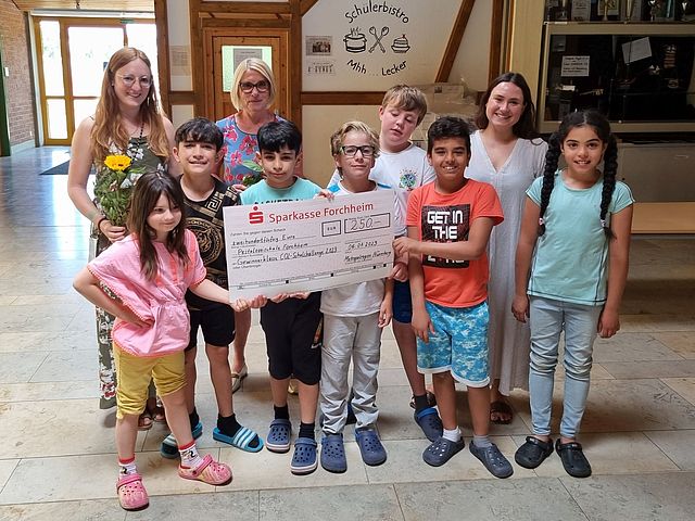 Eine Gruppe von drei Frauen und sieben Schülerinnen und Schüler zeigen stolz einen Gewinnscheck über 250 Euro. Die Gruppe befindet sich in einem Flur eines Gebäudes. Im Hintergrund erkennt man an einer Wand den Schriftzug " Schülerbistro - Mhh---Lecker" und eine Ausstellungsvitrine. 