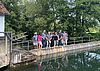 Eine Gruppe aus Frauen und Männern in sommerlicher Kleidung steht auf einem schmalen Wasserwehr über einem grün schimmernden Fluss. Im Hintergrund stehen hohe Bäume.