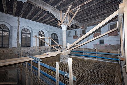 Eine Baustelle in einem großen Raum eines historischen Gebäudes. Am Boden befinden sich Baustahlmatten. In der Mitte des großen Saales steht eine große Steinsäule.