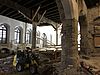 Eine Baustellen-Innenansicht in einer großen historischen Halle mit alter Holzdecke und einer Steinsäule in der Mitte der Halle. Ein kleiner Bagger und Werkzeug sind verstreut in der Halle zu sehen.