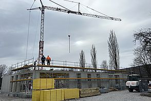 Außenansicht des Rohbaus des Erdgeschosses der neuen Kita mit Baugerüsten und einem Kran im Hintergrund.