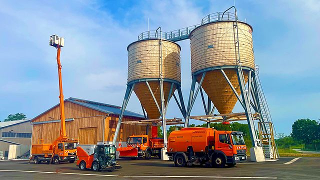 Zwei Hochbehälter für Schüttgut befinden sich rechts neben zwei großen Scheunen. Davor stehen eine große und kleine Kehrmaschine und links ein ausgefahrener Hubwagen.