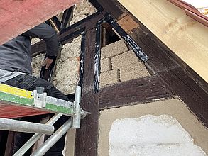 Ein Mauerer sitzt auf einem Baugerüst. Er klebt Fachwerkbalken ab. Das alte Mauerwerk hat teilweise Lücken und wurde wieder teilweise mit Mauersteinen aufgefüllt und teilweise verspachtelt.