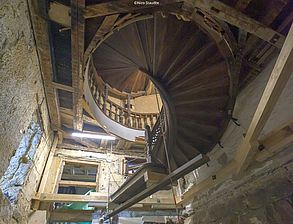 Man sieht eine Wendetreppe aus Holz von unten. Sie ist abgestützt und scheint im Raum zu schweben.
