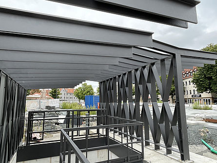 Ein moderner Hochbau aus dunklem Stahl an einem Treppenabgang zu einer Tiefgarage errichtet. Umliegend befindet sich eine Großbaustelle mit Absperrungen und verschiedenem Baumaterial.