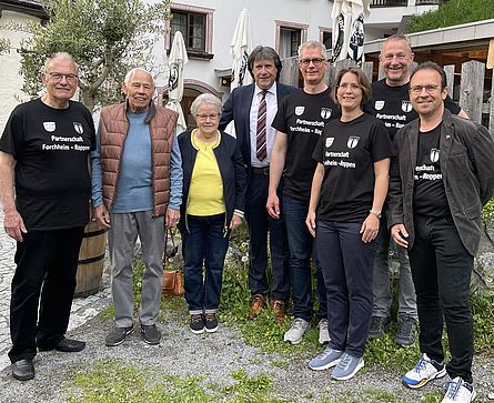 Gruppenfoto mit den Bürgermeistern von Forchheim und Roppen sowie Vertretern des Forchheimer Stadtrates stehen vor einem hellblauen Gebäude. Die Forchheimer Vertreter tragen schwarze T-Shirts mit der Aufschrift " Partnerschaft Forchheim - Roppen". Hinter den Personen sieht man drei weiße, geschlossene Sonnenschirme und einen hohen Holzzaun. In der linken Bildhälfte ragt eine grüne Kübelpflanze auf einem hölzernen Behälter.