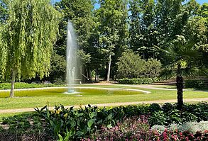 Im Vordergrund ist ein Blumenbeet mit bunten Frühlingsblumen. Im Hintergrund sieht man einen hellgrünen Teich mit Springbrunnen. Links im Bild befindet sich eine Trauerweide und im Hintergrund hohe Bäume. Eine Palme schmückt das Blumenbeet im Vordergrund.